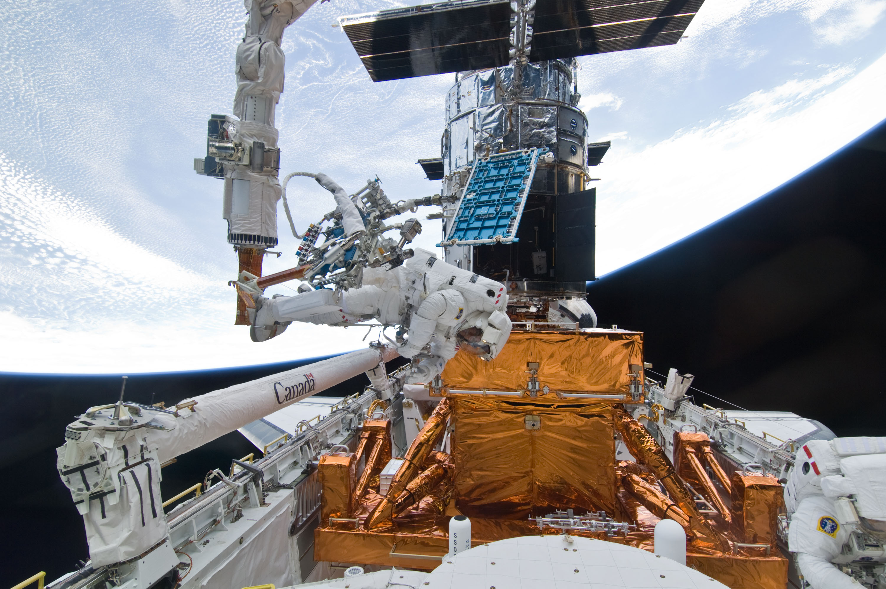 Un astronauta se mueve en el extremo de un brazo robótico del transbordador en el compartimento de carga con numerosas herramientas conectadas mientras otro trabaja en la parte inferior derecha.  Hubble y la Tierra están al fondo.