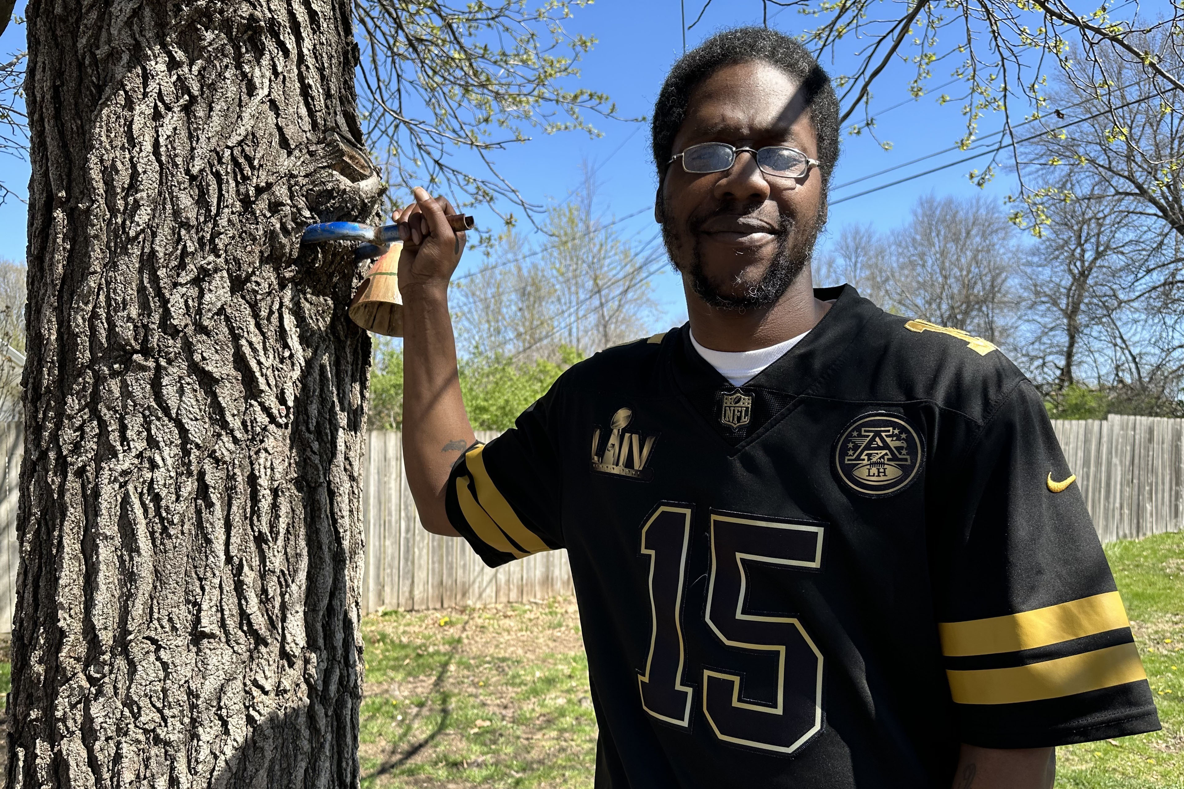 Un hombre con gafas y una camiseta deportiva se encuentra junto a un árbol y posa para una fotografía.