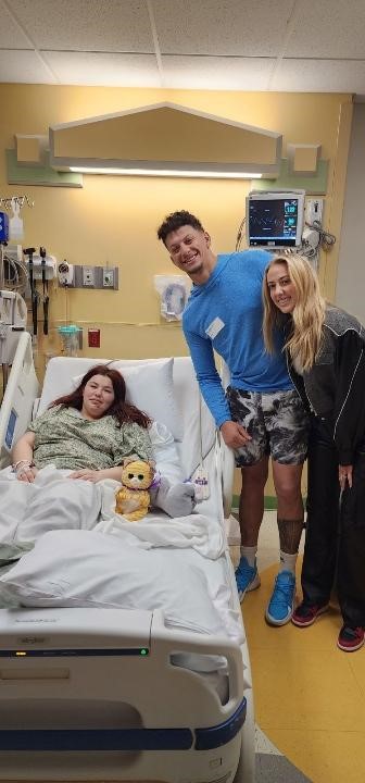 Una foto de una mujer en una cama de hospital.  Un hombre y una mujer están junto a ella, sonriendo.