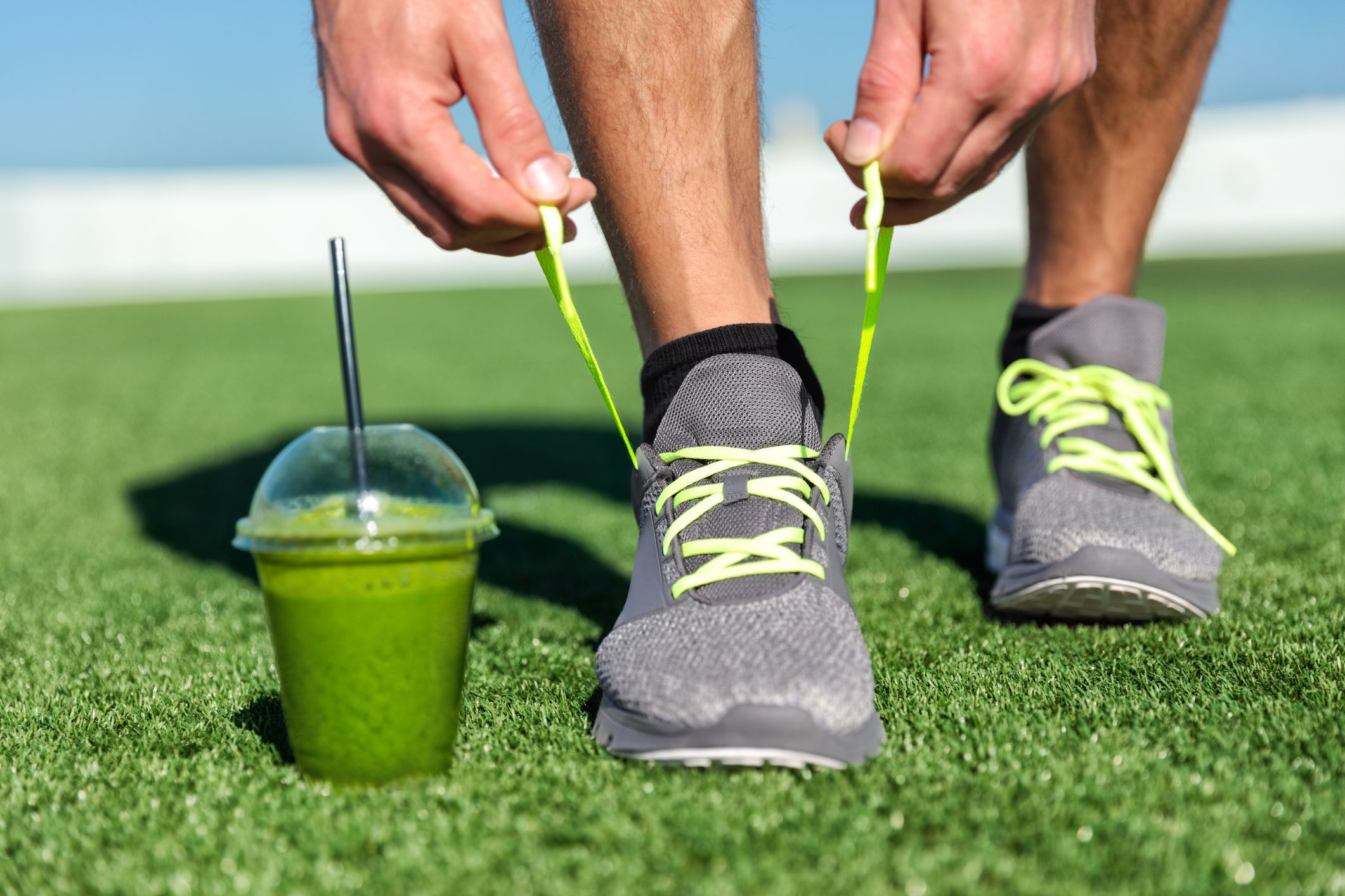 Hombre de fitness con batido verde atando zapatos para correr, corredor atleta con jugo de desintoxicación de vegetales verdes preparándose para correr por la mañana atando cordones de zapatos para correr en fitness de césped y concepto de estilo de vida saludable